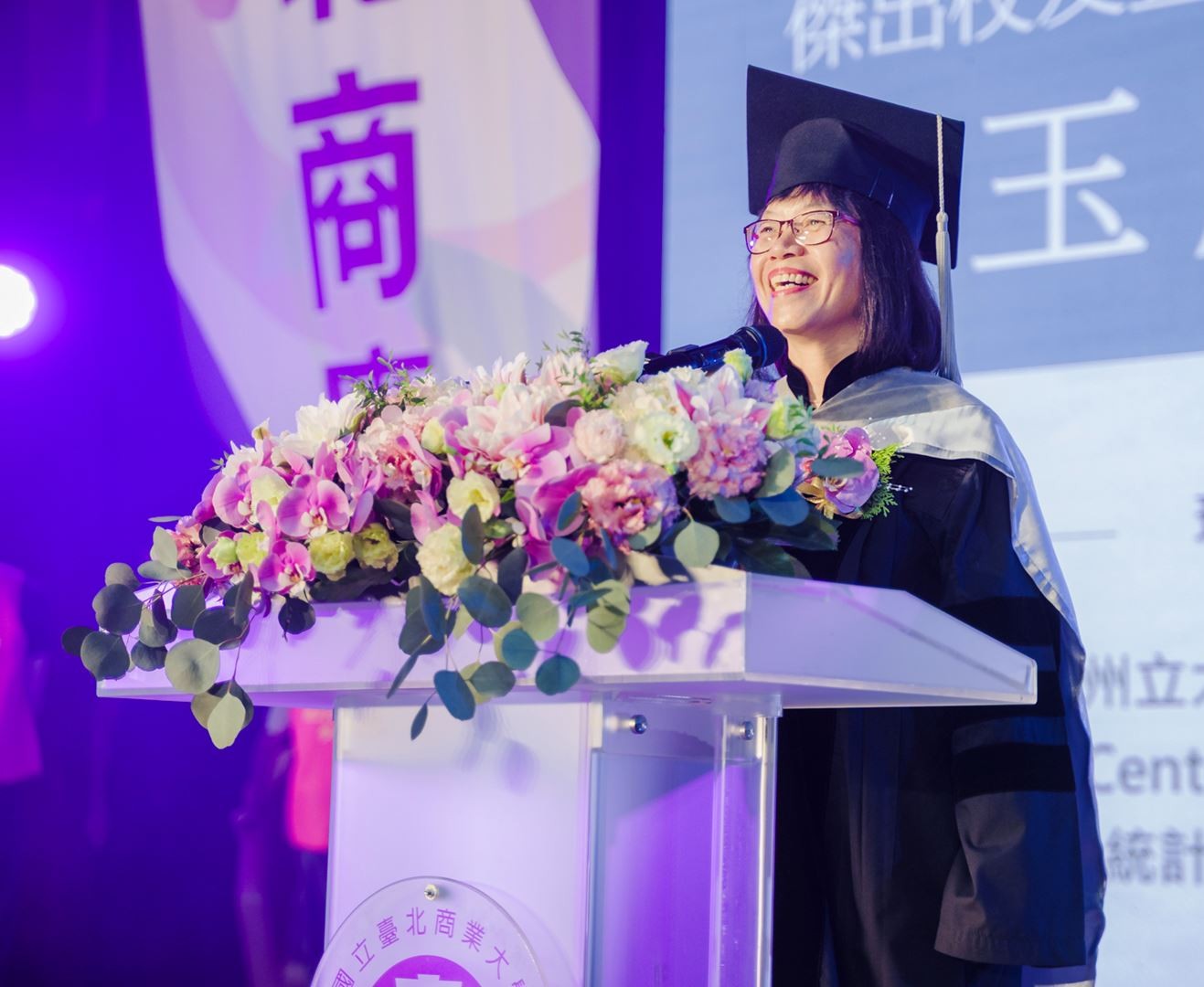 Graduate in cap and gown speaks at a podium decorated with flowers during a commencement ceremony on a purple-lit stage.