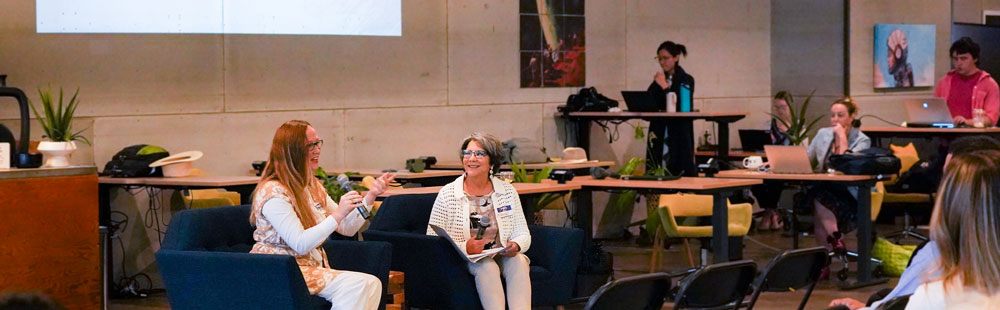 Two people chat on couches in a modern co-working space, with others working at laptops in the background.