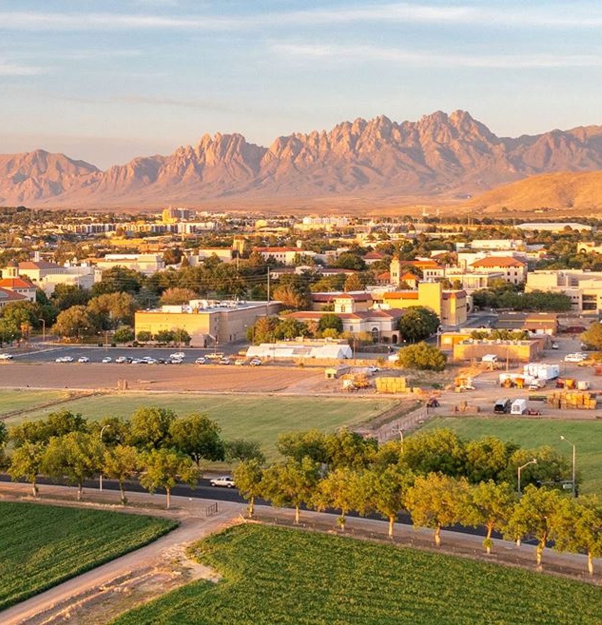 Panoramic view of a small city with red-roofed buildings and tree-lined streets, set against rugged mountains at sunset.