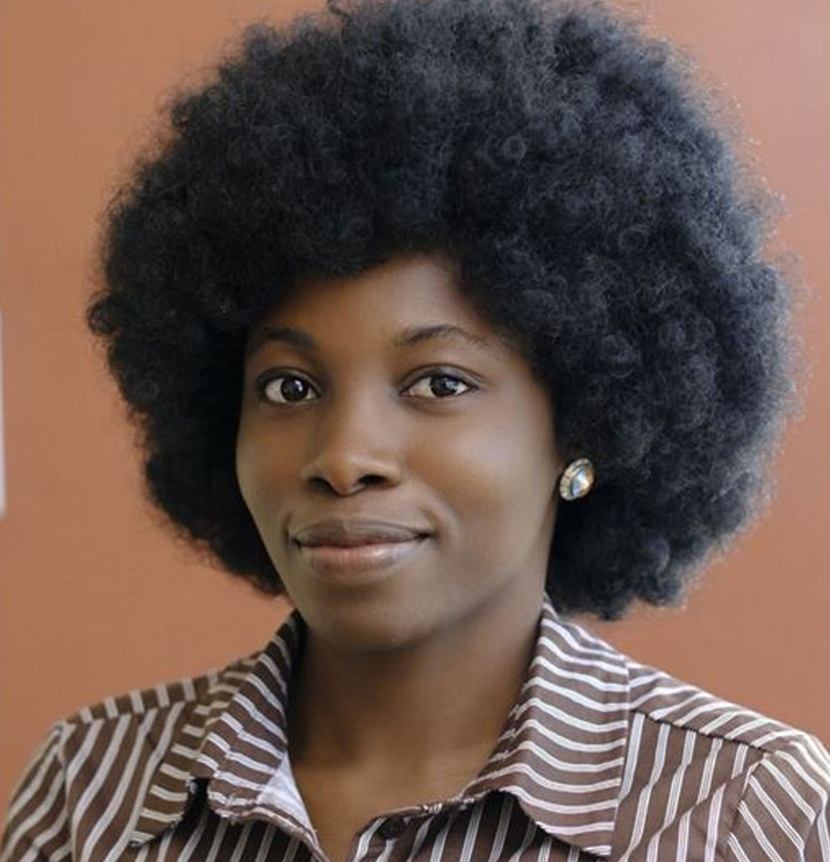 Head-and-shoulders portrait of a woman with a natural afro hairstyle, wearing stud earrings and a striped collared shirt against a plain background.